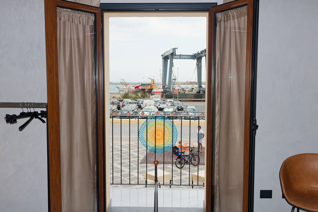 La portafinestra della camera spalancata sul balconcino: il centrino all'uncinetto legato alla ringhiera e, oltre la strada, le barche e le gru del porto