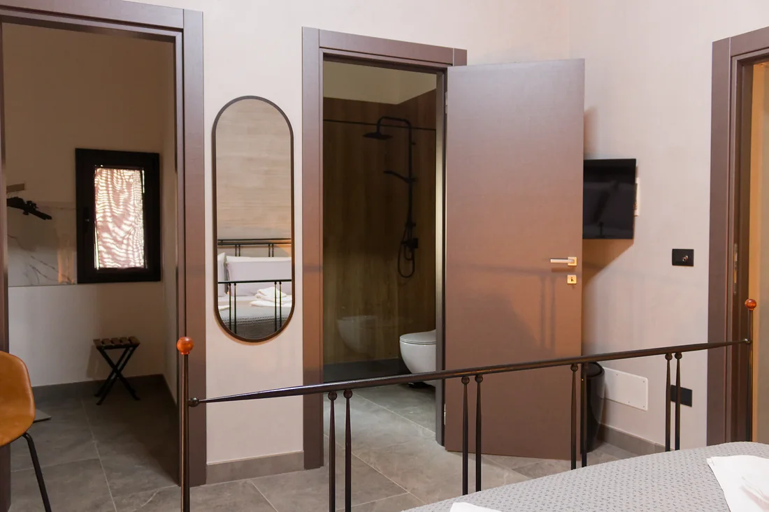 La camera verso il bagno: la porta aperta sulla doccia, lo specchio ovale accanto e il televisore alla parete