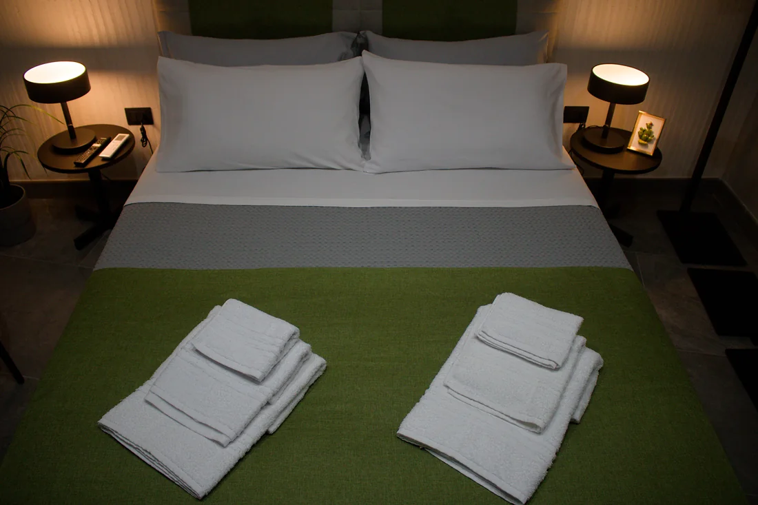 Il letto visto dall'alto, con la coperta verde, gli asciugamani piegati e le due lampade accese sui comodini