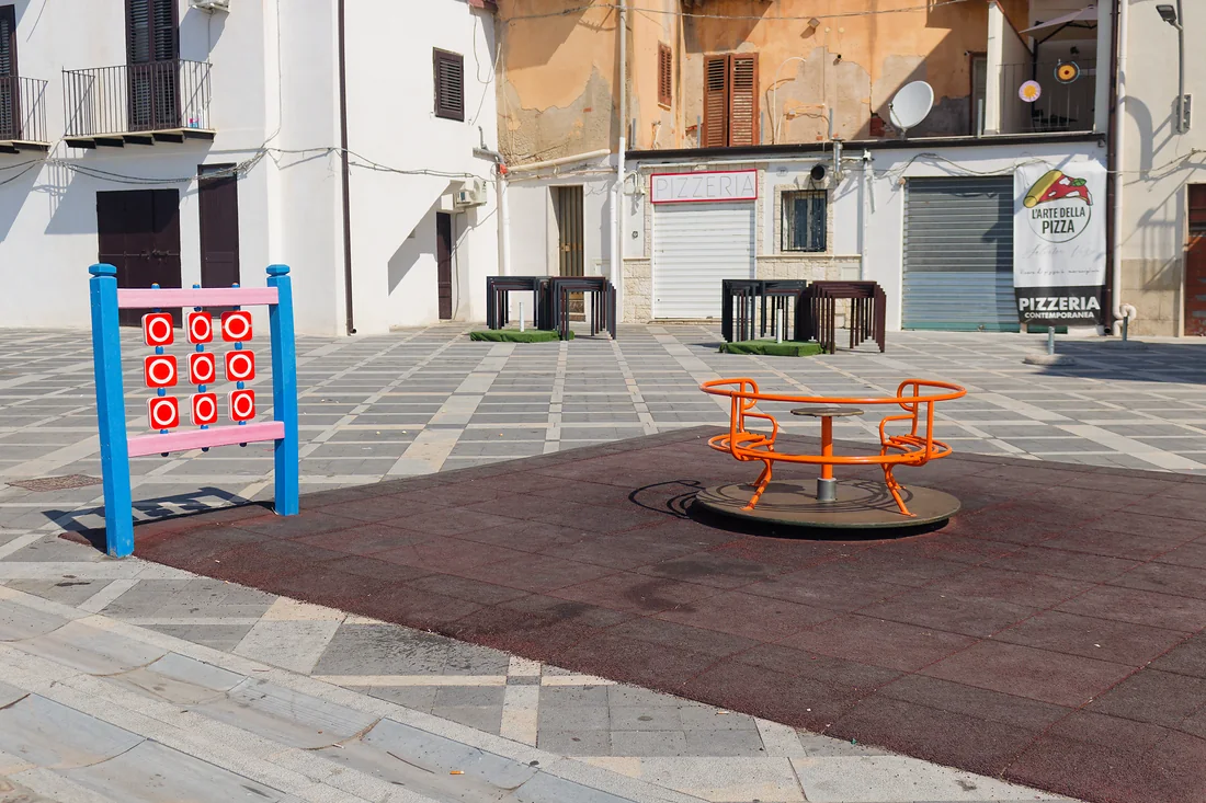 La piazzetta pavimentata a scacchi, con l'area giochi: il gioco del tris a colonnine colorate, la giostrina arancione e, in fondo, le saracinesche di una pizzeria