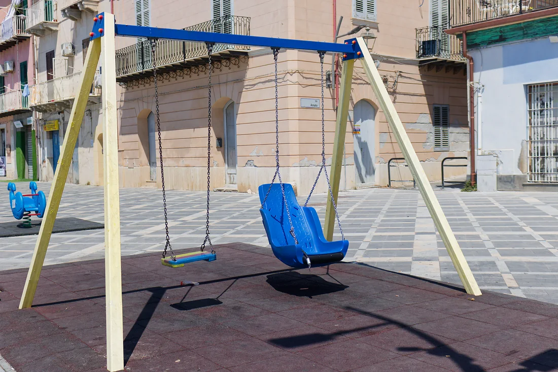 L'altalena dell'area giochi, con un seggiolino azzurro e uno a tavoletta, davanti a un palazzo rosa d'angolo; a sinistra una balena a molla azzurra