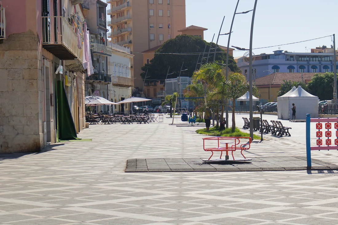 La piazza sul lungomare, pavimentata a losanghe: la giostrina rossa dell'area giochi, le panchine, le palme e i tavolini di un bar in fondo