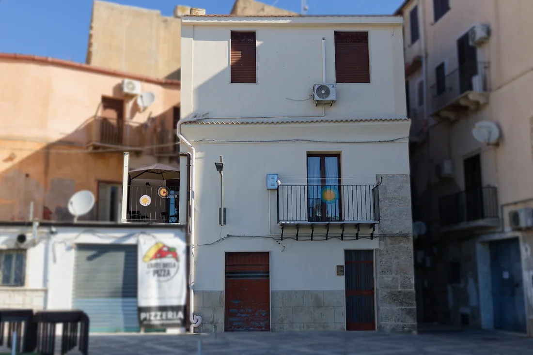 La facciata vista dalla piazzetta: la casa bianca su tre livelli, il balconcino con il sole di ceramica e il portone; a sinistra l'insegna della pizzeria accanto